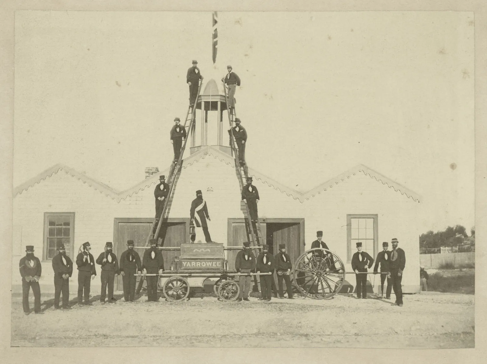 About twenty-five men in uniform and caps lined up in front of a gabled hall with fretwork bargeboards, around a fire engine lettered YARROWEE and a hose reel, with a tall pyramid of ladders over the engine and more men posed on its rungs.