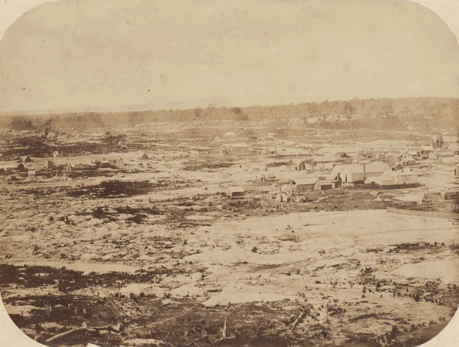 A wide view across worked-over diggings: heaps, holes and pools of water in the foreground, a line of buildings and trees along the far rise.