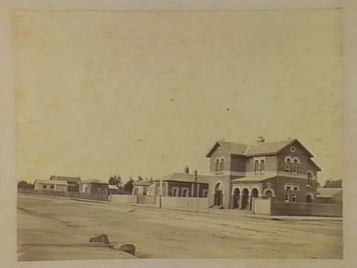 A wide unmade road; on the right a two-storey brick building with arched openings, then a row of single-storey brick and timber buildings behind fences.