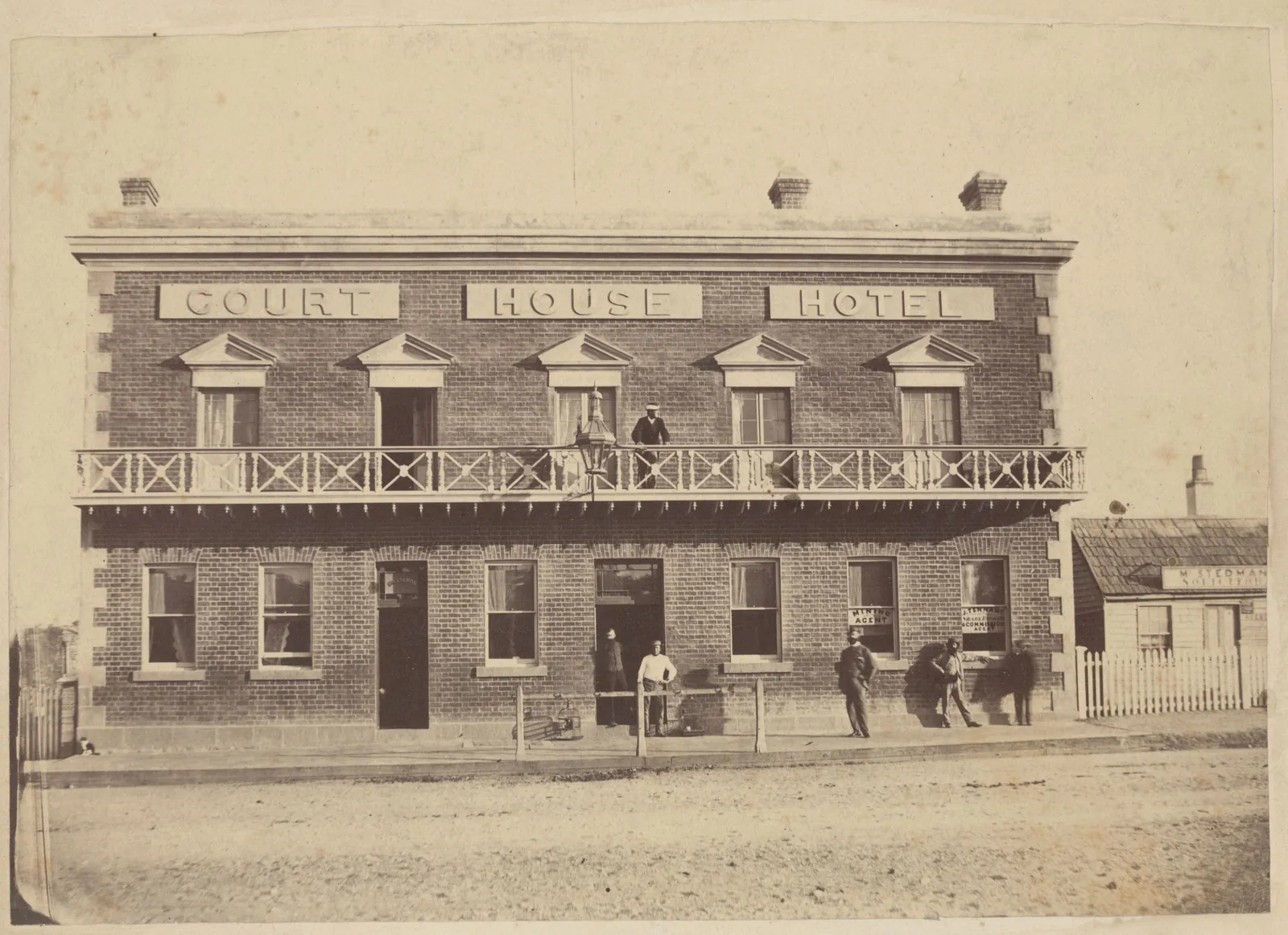 A two-storey brick hotel lettered COURT HOUSE HOTEL across its parapet, a timber balcony with cross-braced railings, people standing on the footpath and one on the balcony, a weatherboard cottage beside it.