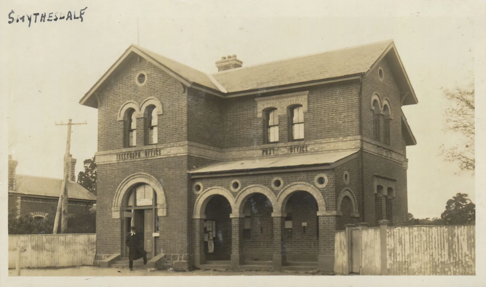 A two-storey red-brick post office with an arched loggia of three bays on the ground floor and POST OFFICE lettered across its front, a telegraph pole and a picket fence beside it.