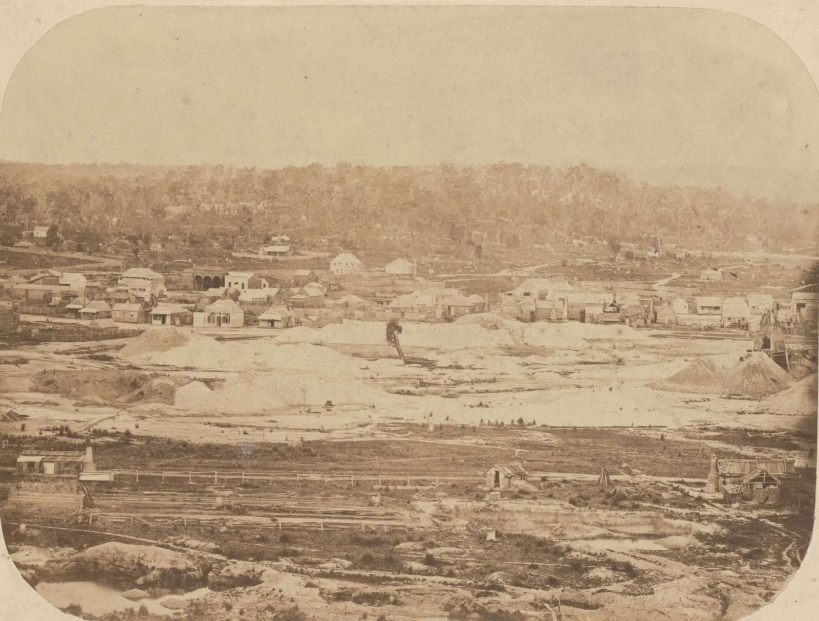 The town seen across open diggings: a long line of buildings along the far side of the worked ground, with pale heaps of washdirt and a figure standing among them.
